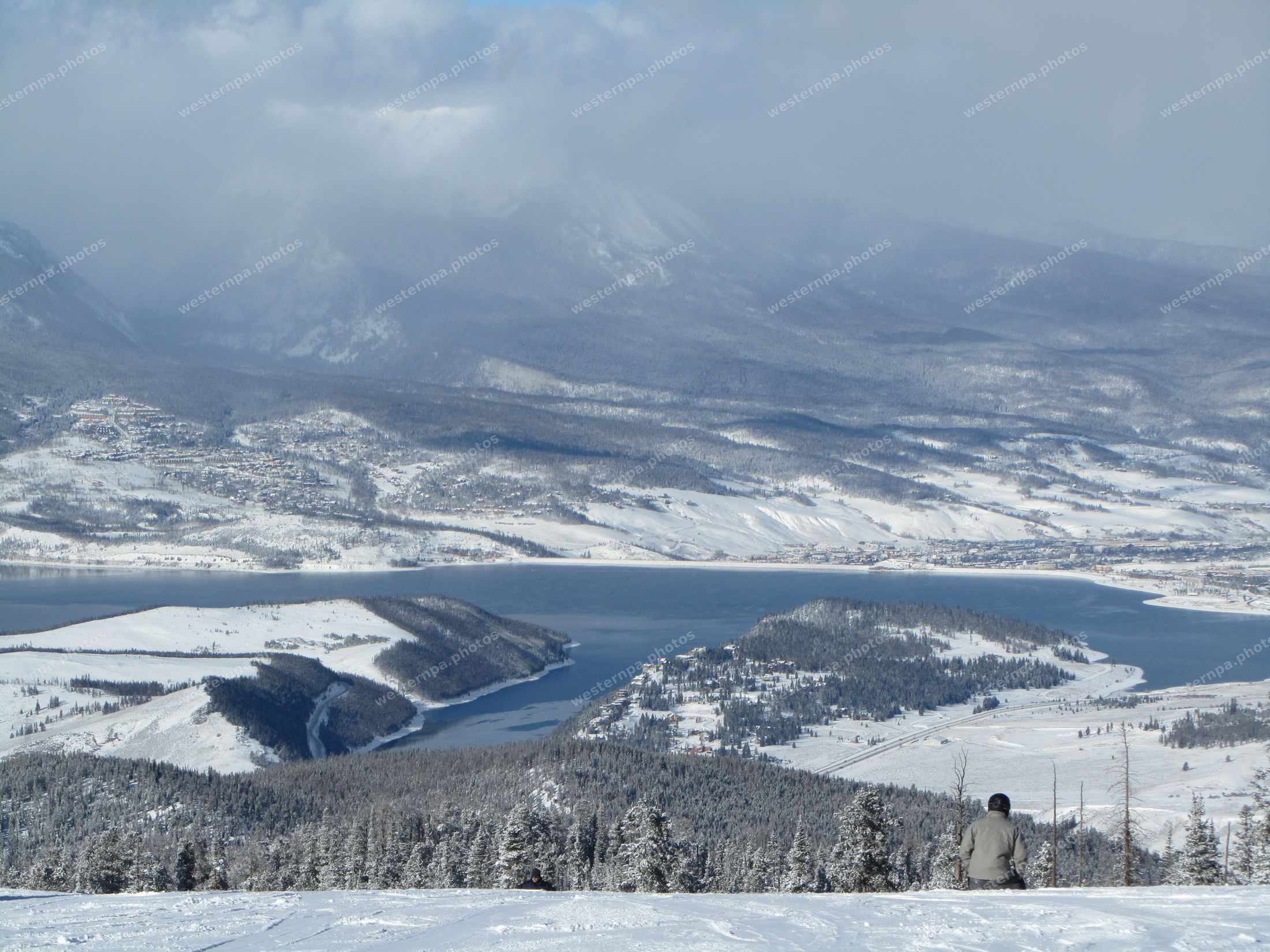Skier from behind looking down on a partly frozen mountain reservoir ringed by snowy peaks