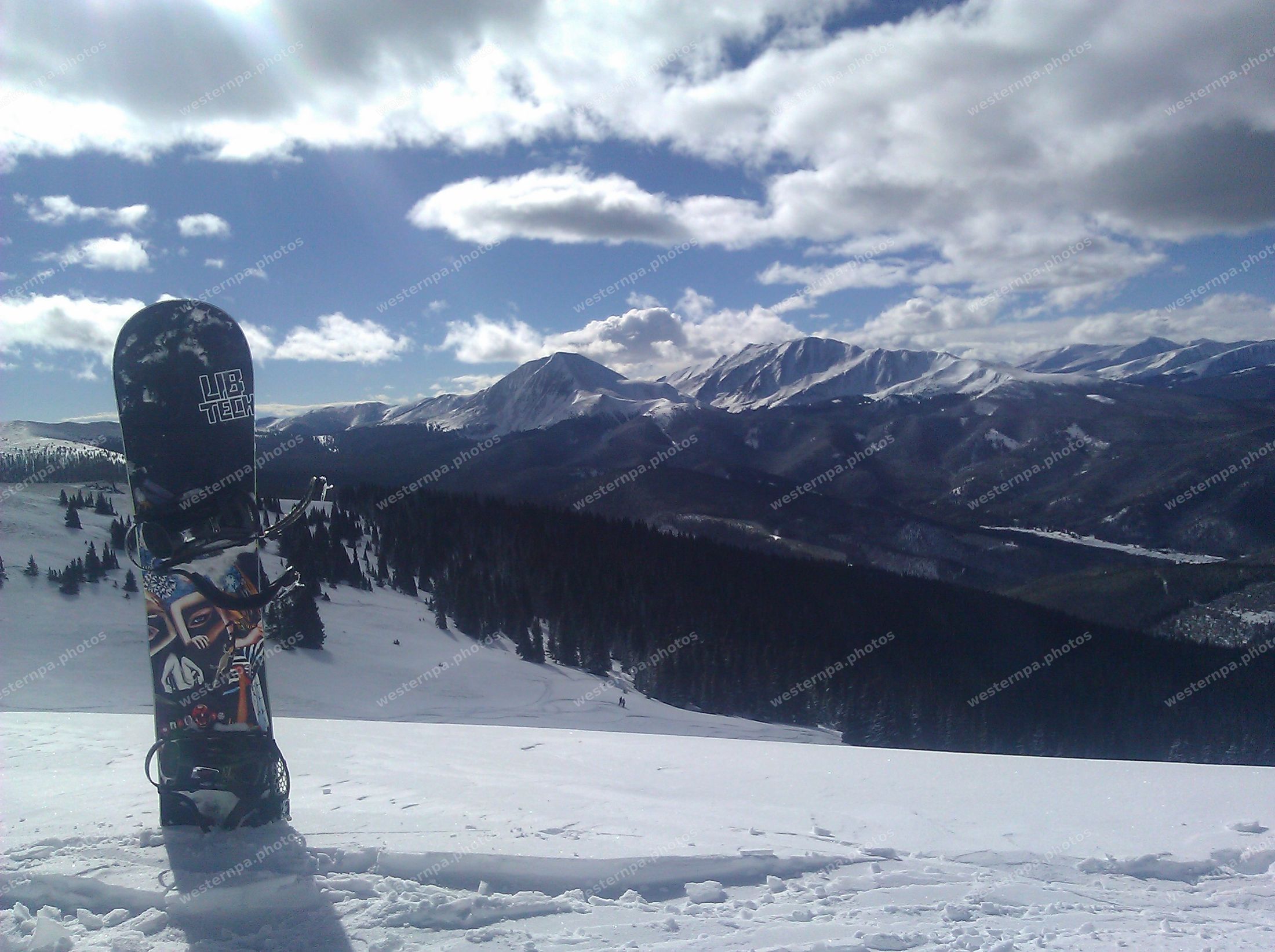 Snowboard standing upright in snow with sunlit winter peaks and forest behind