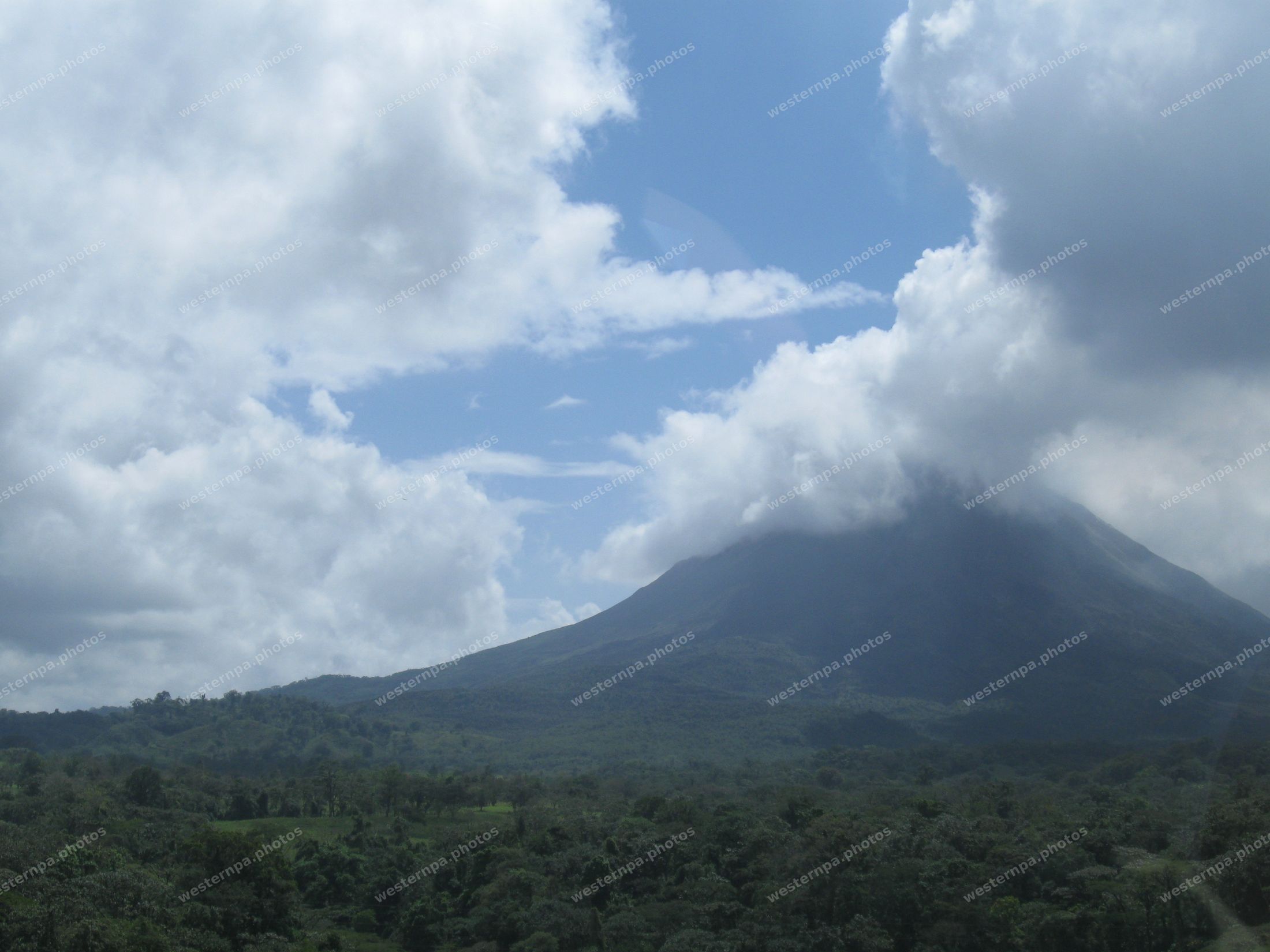 Volcanic cone wrapped in clouds rising above dense green rainforest under a blue sky